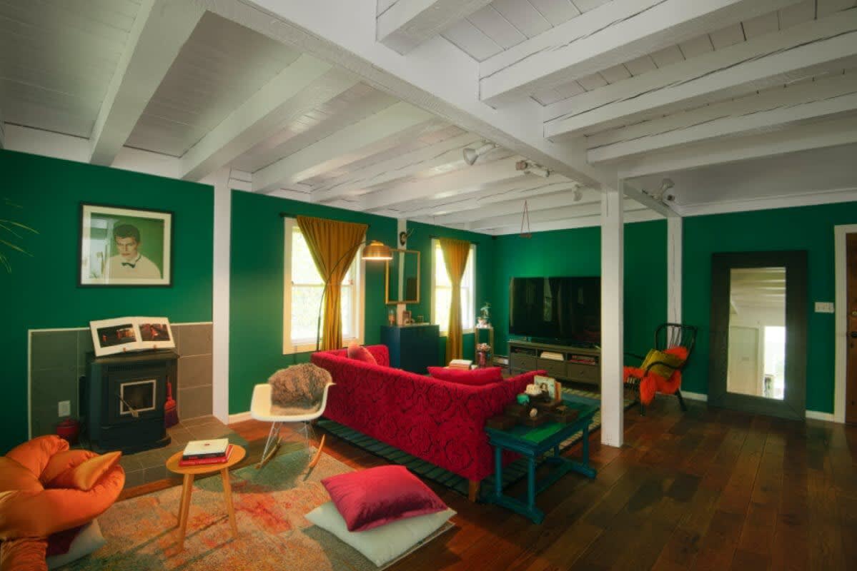 Vibrant living room with emerald green walls and exposed white ceiling beams, featuring a red patterned sofa and a wood-burning stove. Yellow curtains frame windows overlooking the outdoors.