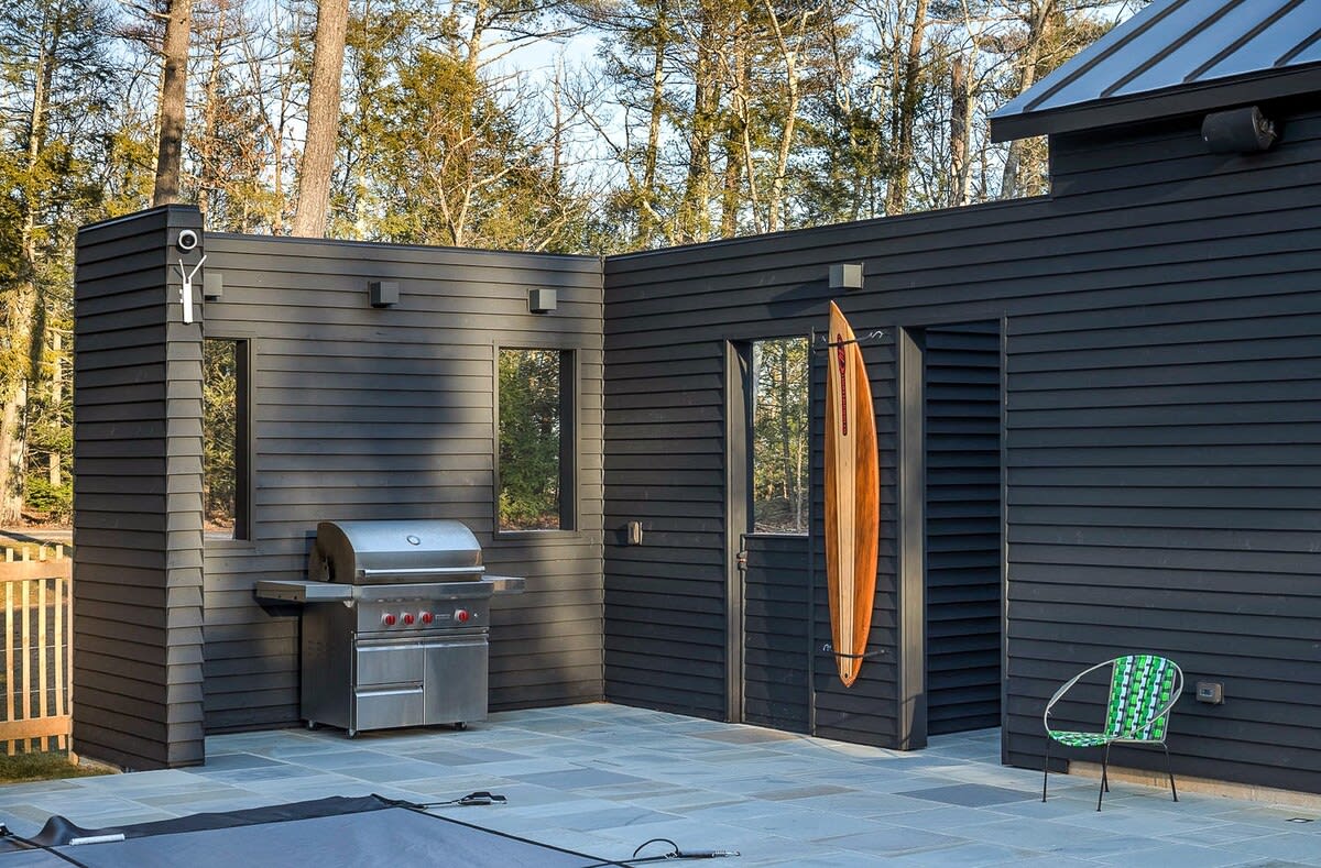 Exterior view of a black horizontal-slat sided building with a stainless steel BBQ grill on a stone patio. A wooden surfboard is mounted on the wall, and a green wire chair adds a pop of color.
