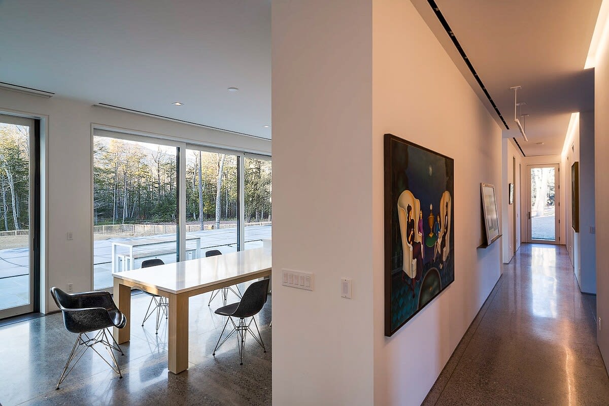 A bright dining area featuring a long white table and black wire-frame chairs, positioned in front of large windows overlooking a natural, wooded landscape. A well-lit hallway with artwork extends to the right.