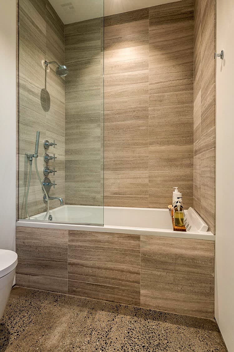A contemporary shower-over-tub combination with light brown, horizontally-tiled walls and a clear glass partition. The white bathtub and modern shower fixtures are visible, set against a polished concrete floor.