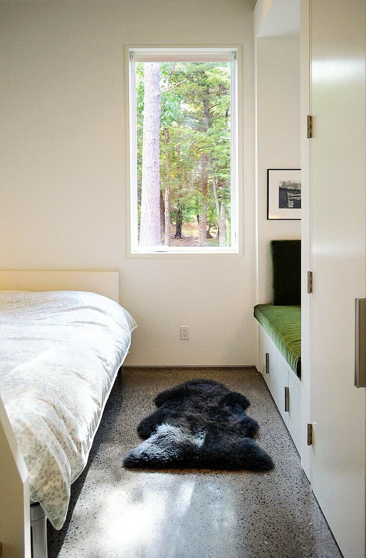 Minimalist bedroom with a white bed, concrete flooring, and a large window offering views of lush trees. A dark sheepskin rug adds texture, and a built-in green cushioned bench provides seating.