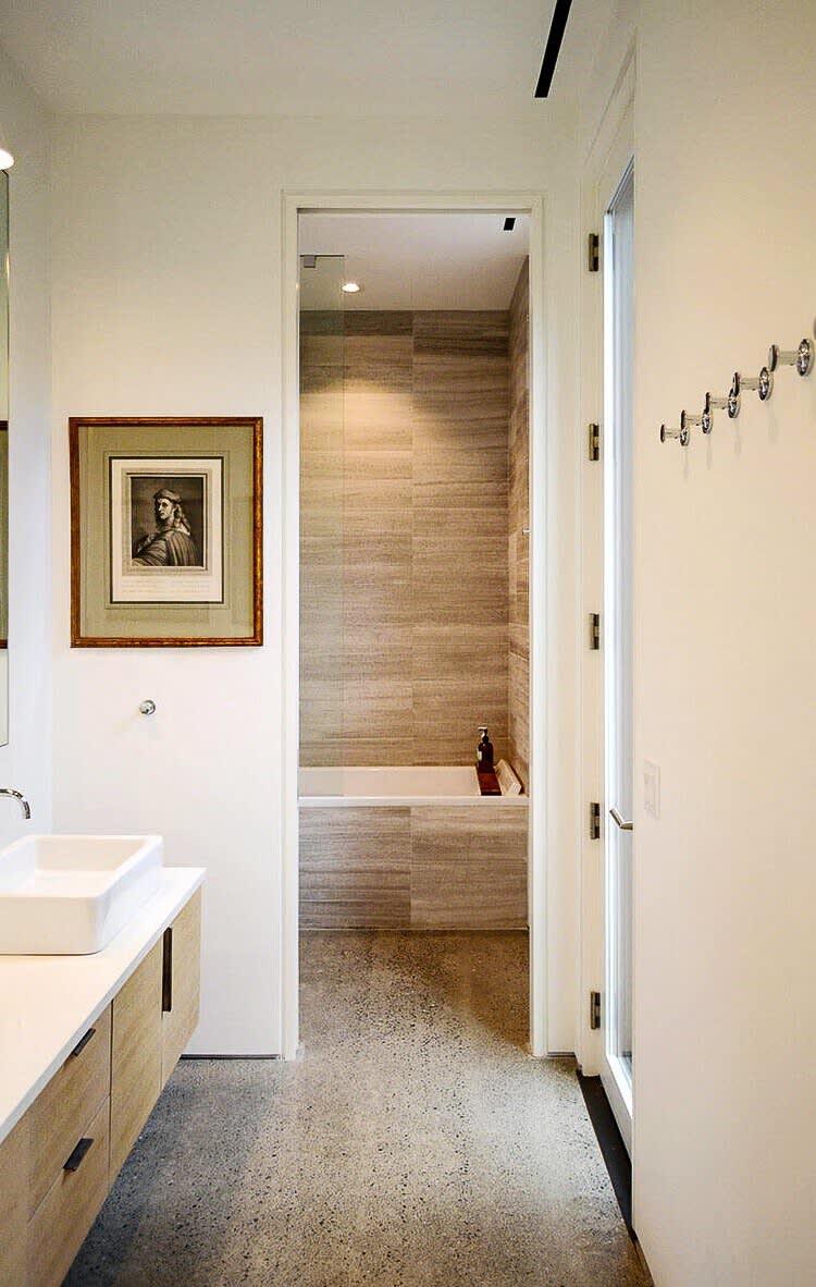 Modern bathroom featuring a white floating vanity with a vessel sink, concrete flooring, and a separate alcove with a built-in bathtub and tiled walls. A framed artwork hangs on the wall, and a door leads to another room.