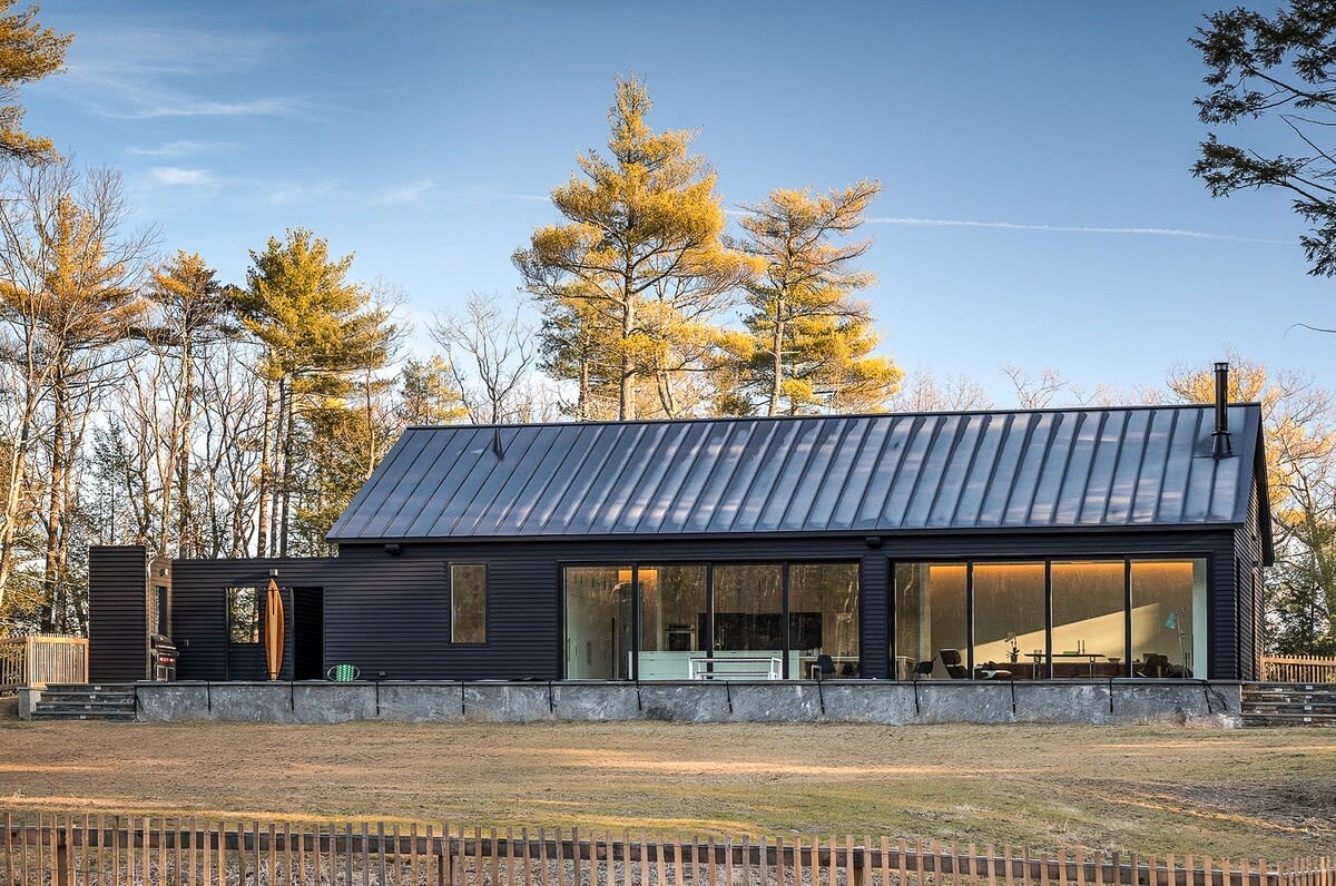 Modern black-clad house with a dark metal roof, featuring large windows and set against a backdrop of tall trees and a grassy yard. The contemporary design blends with the natural surroundings under a clear sky.