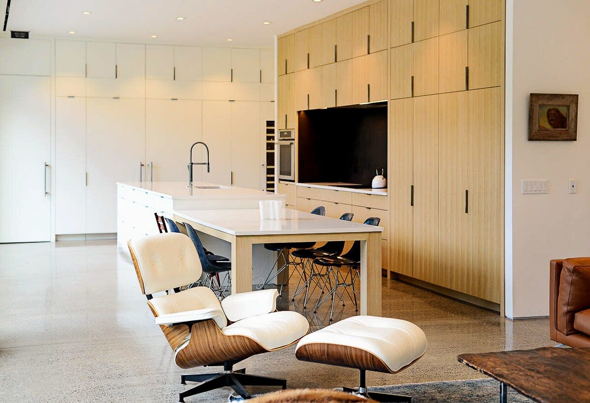 An open-concept living space showcasing a white Eames lounge chair and a brown leather sofa, centered around a large wooden coffee table. The adjacent modern kitchen features light wood and white cabinetry.