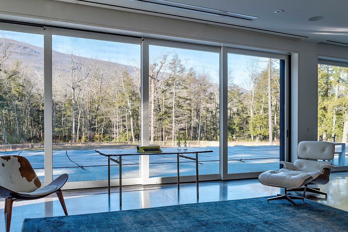Bright living area with polished concrete floors and expansive floor-to-ceiling sliding glass doors offering panoramic views of a winter forest and distant mountains. An Eames lounge chair and a cowhide chair furnish the space.