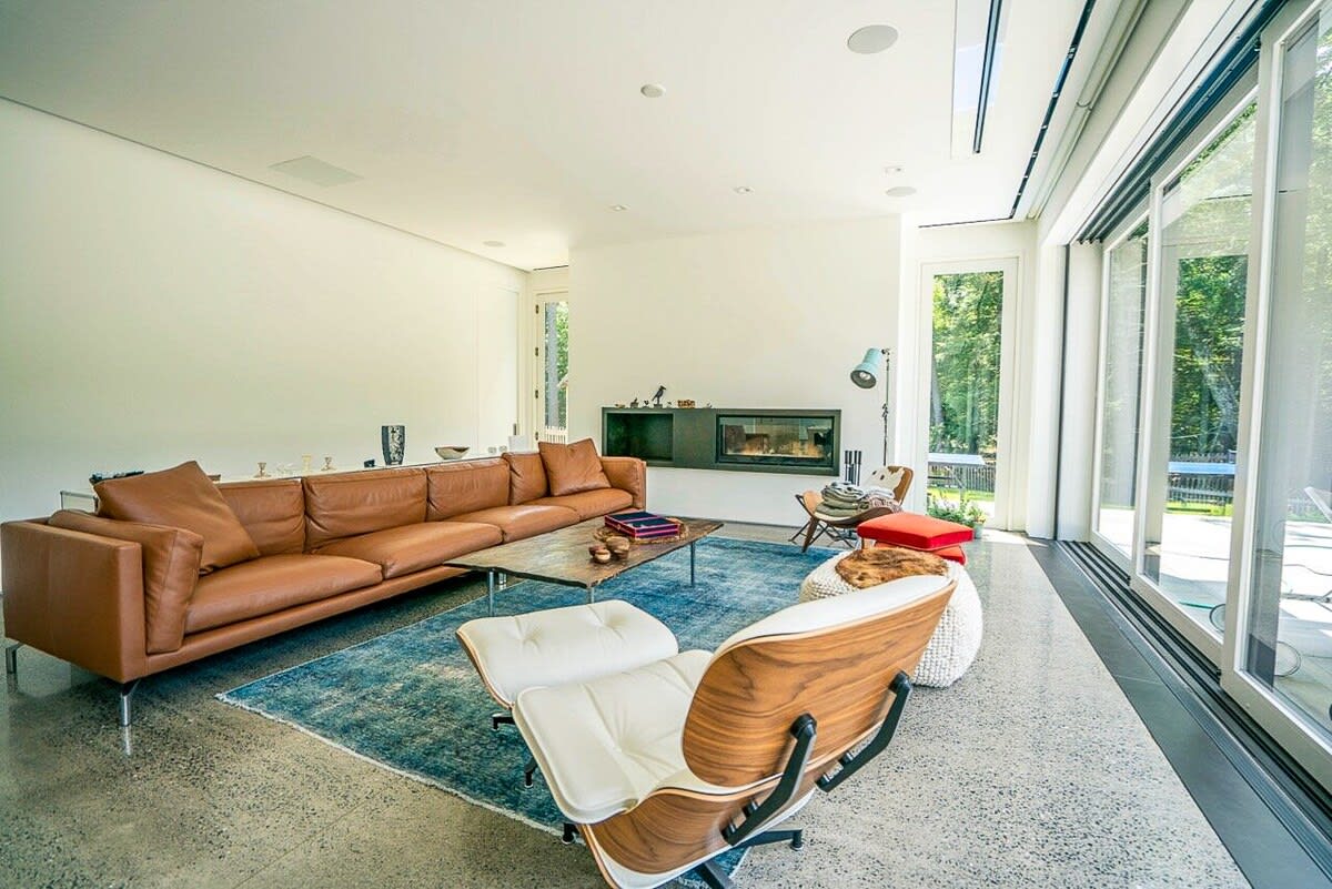 Spacious living room featuring a long brown leather sofa, a blue area rug, and a modern fireplace built into a dark wall unit. Polished concrete floors extend to large sliding glass doors overlooking the pool and forest.