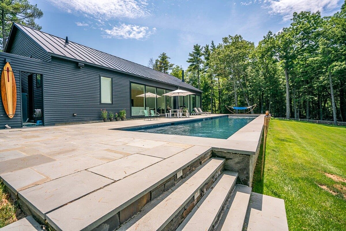 Side view of the modern black house with a dark metal roof, featuring a long rectangular swimming pool and a stone patio. A wooden surfboard hangs near the entrance, and steps lead down to a grassy yard.