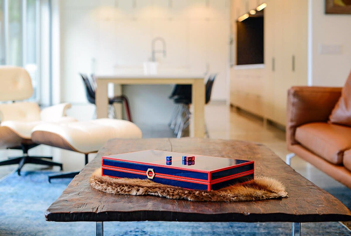 A modern living room with a rustic wooden coffee table, topped with a blue and red decorative box and dice. A white Eames lounge chair sits to the left, with an open-concept kitchen visible in the background.