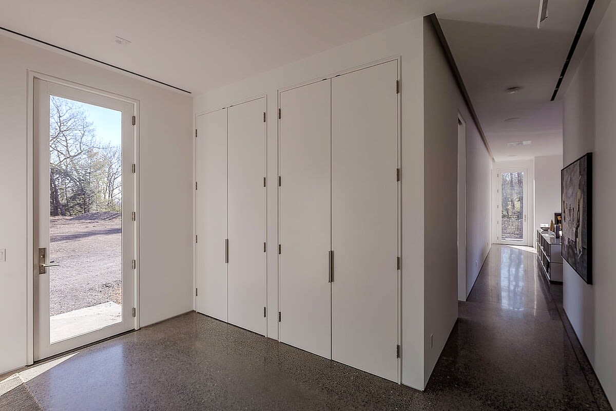 Bright hallway with polished concrete floors, multiple white flush-panel closet doors, and a glass-paned exterior door leading to an outdoor area. The space features clean lines and minimalist design.