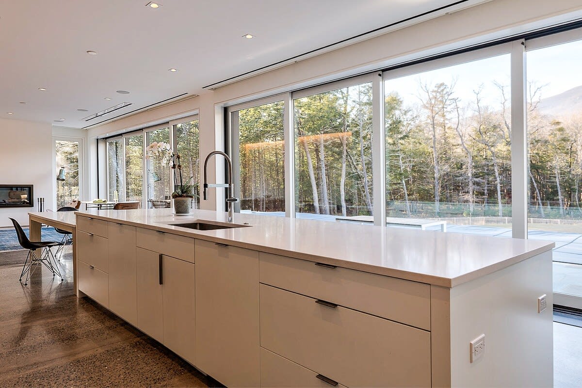 A sleek, modern kitchen featuring a long white island with an integrated sink and contemporary faucet. Light-colored cabinetry lines the walls, and expansive sliding glass doors offer views of a dense wooded area.