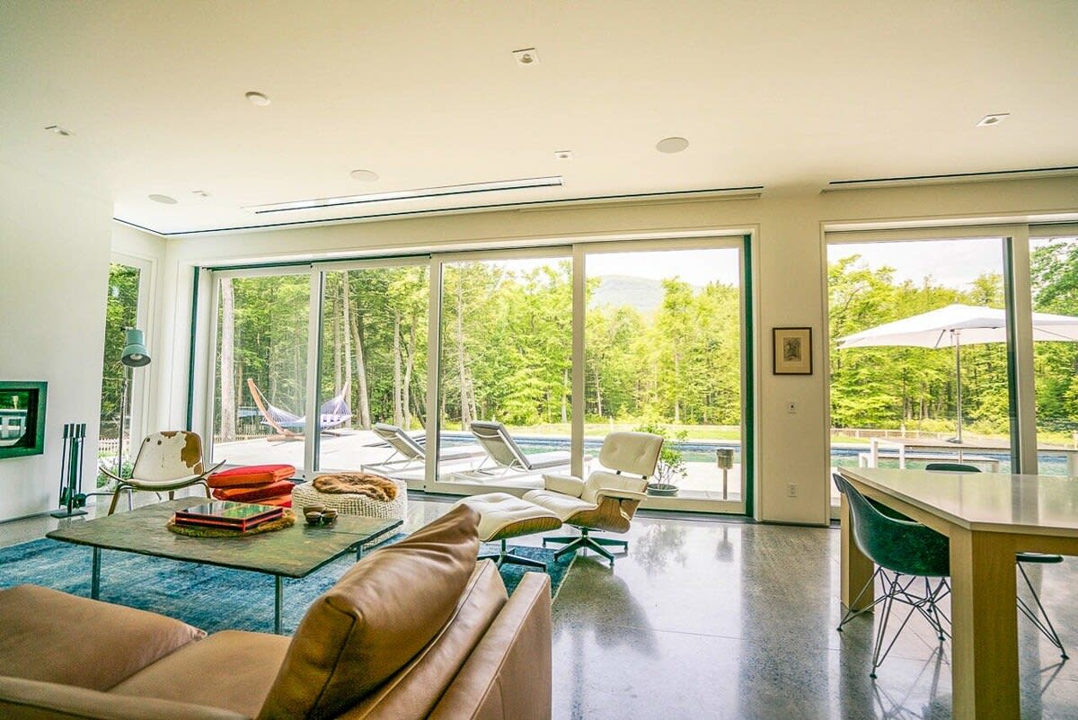 Bright living room with polished concrete floors and a brown leather sofa, facing expansive floor-to-ceiling glass doors that open to an outdoor pool and a lush green forest. An Eames lounge chair is also visible.