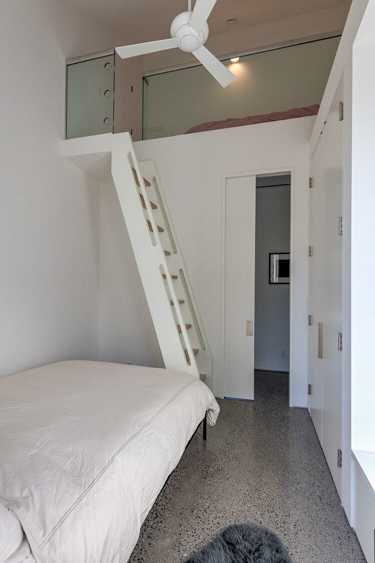 A compact bedroom featuring a bed on the main floor and a white wooden ladder leading to a lofted sleeping area with a glass railing. The room has clean white walls, a ceiling fan, and a polished concrete floor.