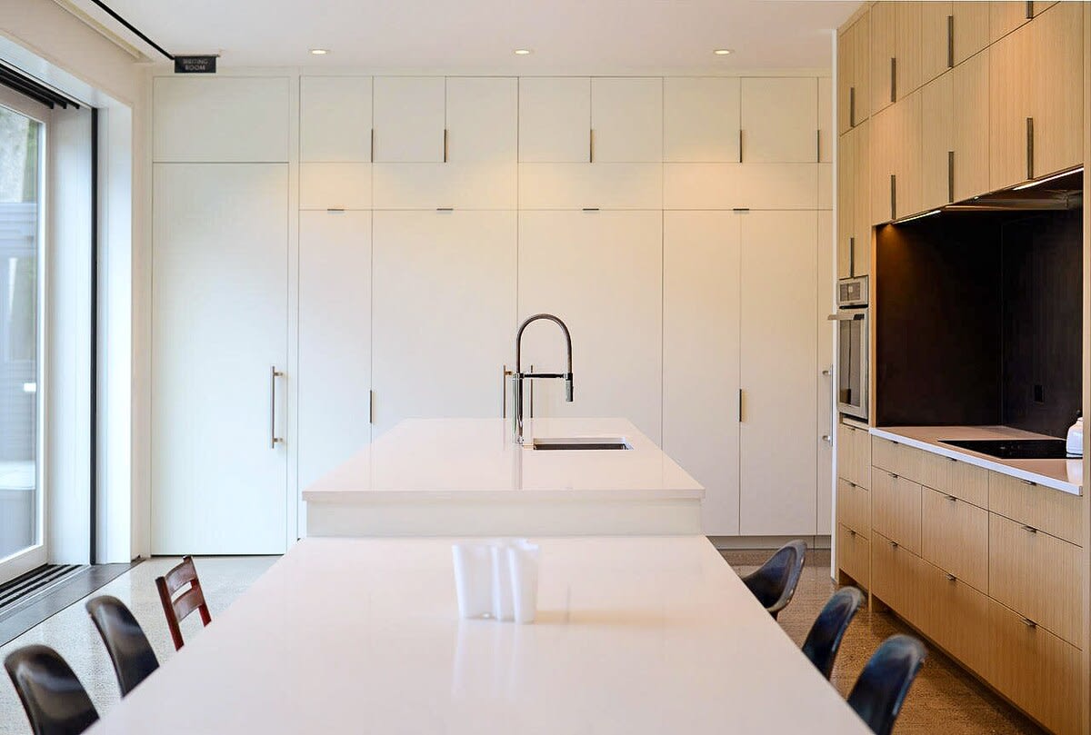 A contemporary kitchen with a long white island extending into a dining table, surrounded by light wood and white flat-panel cabinetry. A large sink and modern faucet are prominent, with a wall of white cabinets on the left.