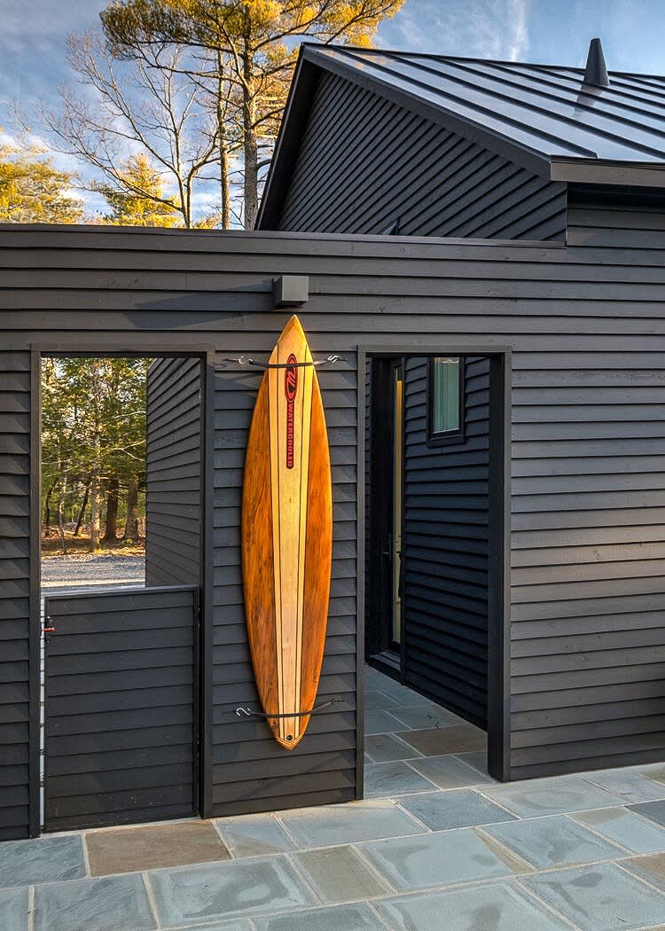 Close-up exterior view of a black horizontal-slat sided building with an open doorway, revealing a glimpse of the interior. A decorative wooden surfboard is prominently displayed on the exterior wall, next to a gate.