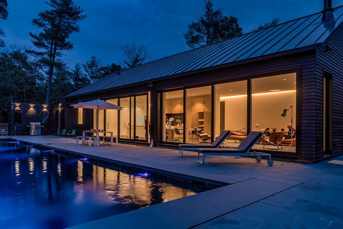 Modern black-clad house with a dark metal roof, featuring expansive floor-to-ceiling windows that illuminate the interior living spaces at dusk. A rectangular swimming pool with blue lighting is surrounded by a stone patio with lounge chairs and an outdoor dining area.