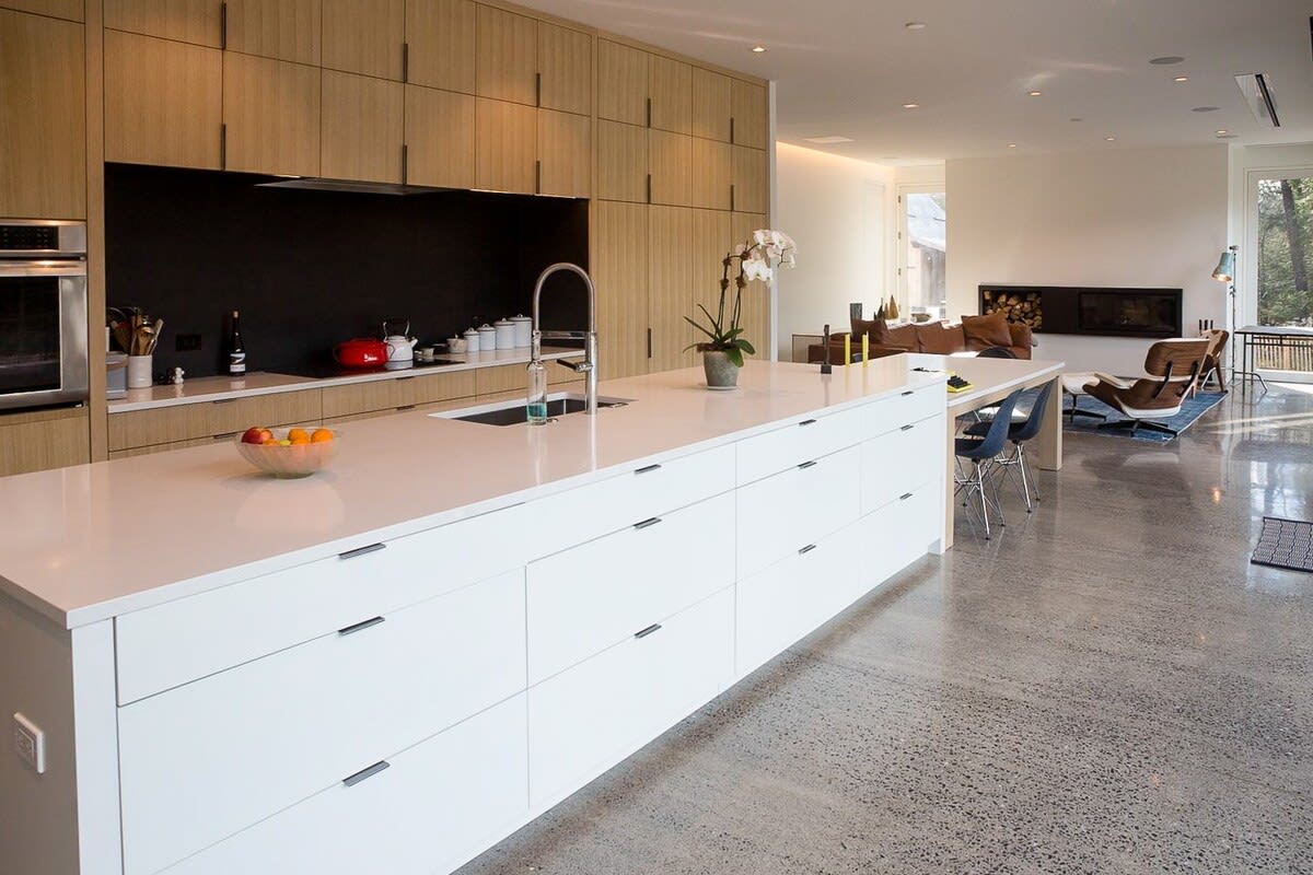 Modern kitchen with a large white island featuring ample storage and a sink, complemented by light wood-paneled cabinetry and a black backsplash. Polished concrete floors extend throughout the open space.