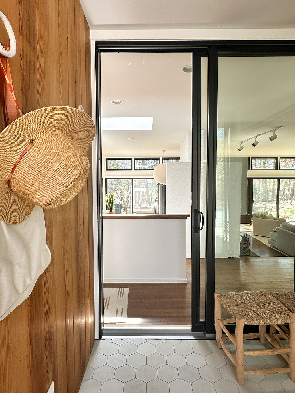 A bright entryway featuring hexagonal white floor tiles and warm wood-paneled walls, with a straw hat hanging on a hook and a black-framed sliding glass door opening into an open-concept living space.