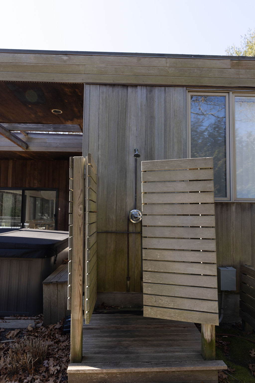 An outdoor shower with wooden slatted privacy screens, located on a wooden deck next to the house's weathered wooden siding. A hot tub is partially visible in the background.