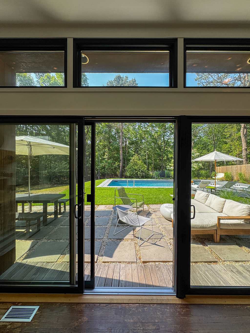 View from inside through large black-framed sliding glass doors to an outdoor patio with modern seating, leading to a lush green lawn and a swimming pool surrounded by mature trees.