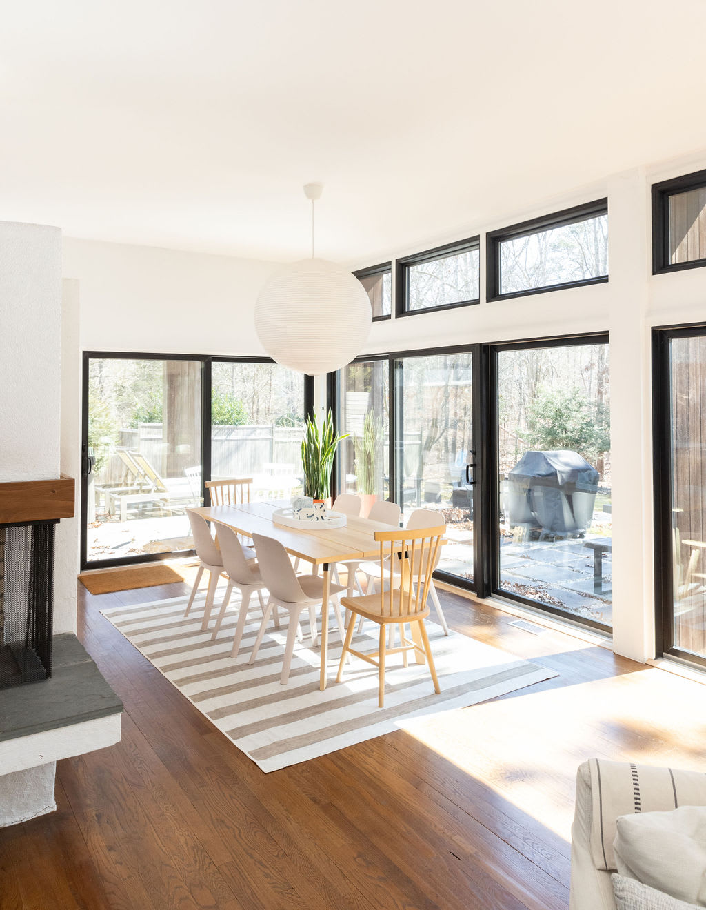 A sunlit dining area featuring a long light wood dining table with a mix of modern and classic chairs, set on a striped rug, with floor-to-ceiling windows offering expansive views of the outdoors.
