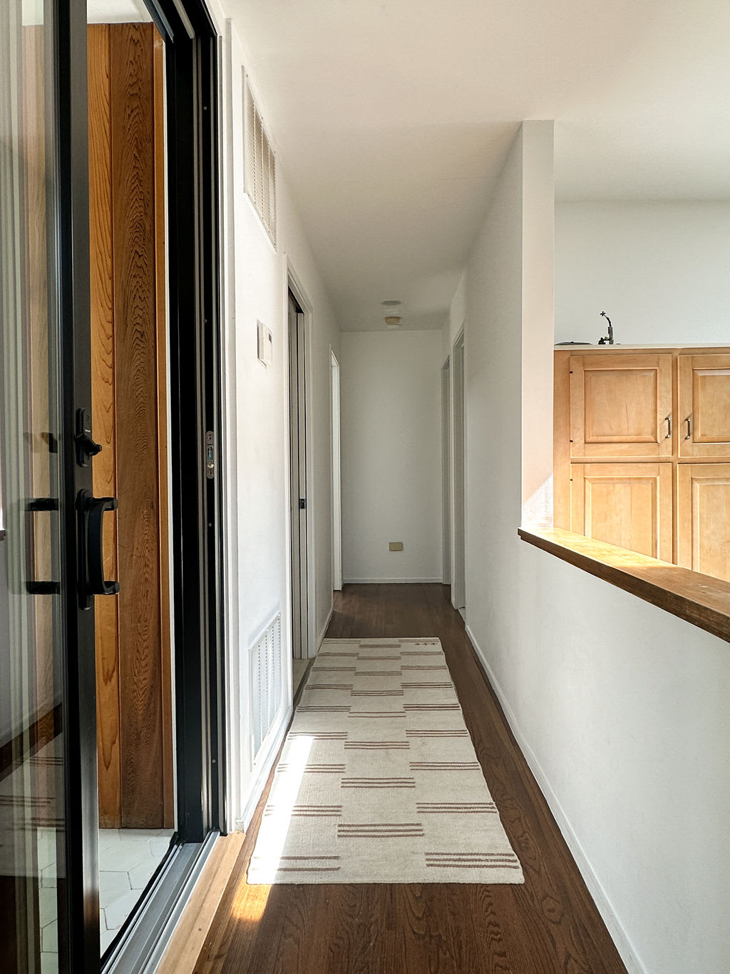 Narrow hallway with dark wood floors and white walls, featuring a striped runner rug. A large sliding glass door is on the left, and a partial view of kitchen cabinets is visible in the distance on the right.