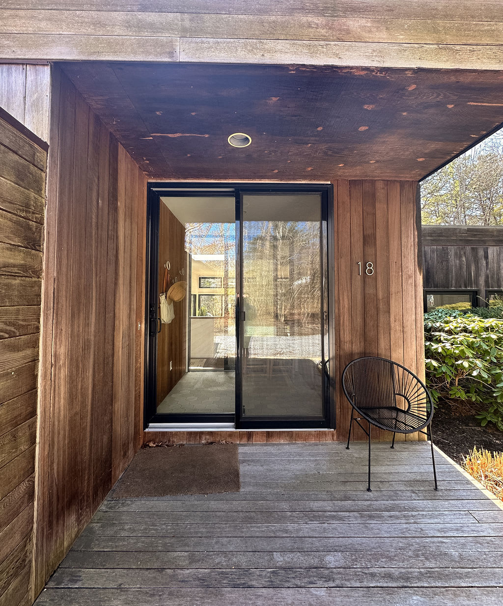 A slightly wider view of the front entrance, showcasing the modern wooden architecture with a covered porch area and a large glass sliding door. A black wire chair sits on the wooden deck, with the house number "18" visible.