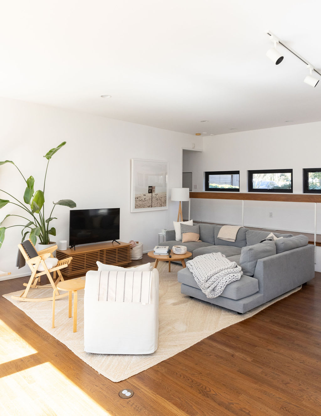 A bright living room with a grey sectional sofa and a light-colored area rug, featuring a wooden media console beneath a wall-mounted TV, with ample natural light streaming in from large windows.
