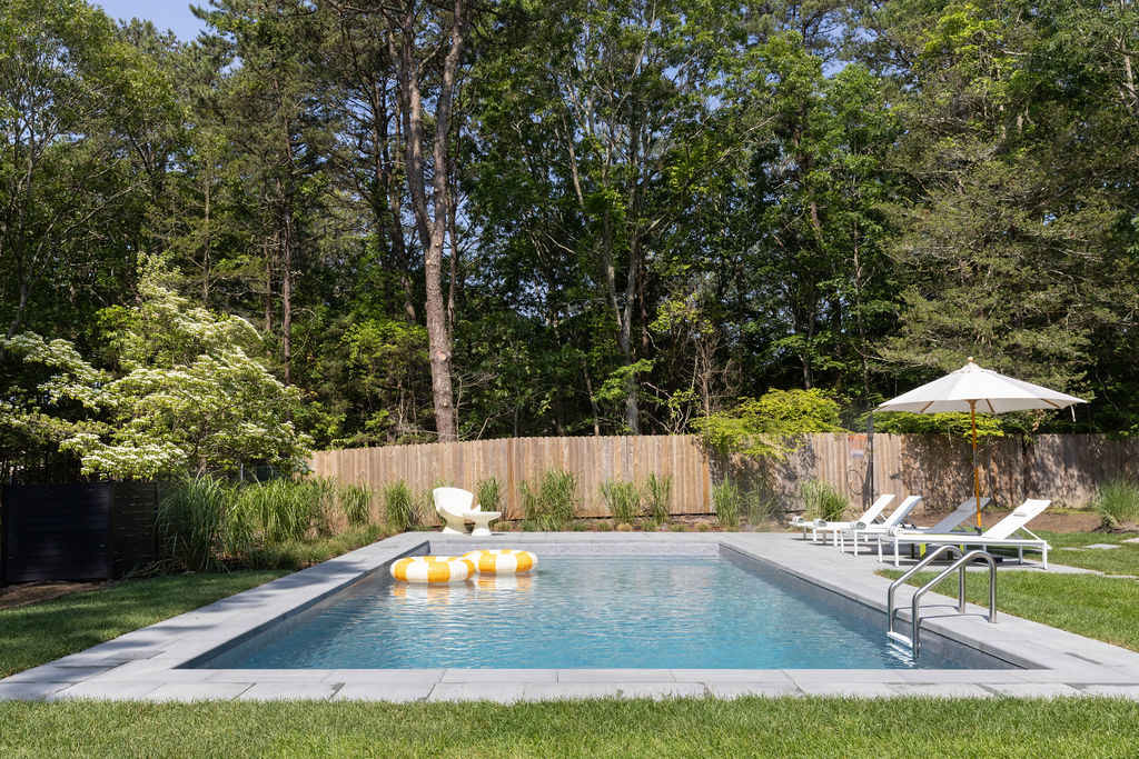 Rectangular swimming pool with a light gray paved surround, two floating rings, and white lounge chairs. The pool is set in a well-maintained green lawn, bordered by a wooden fence and mature trees.
