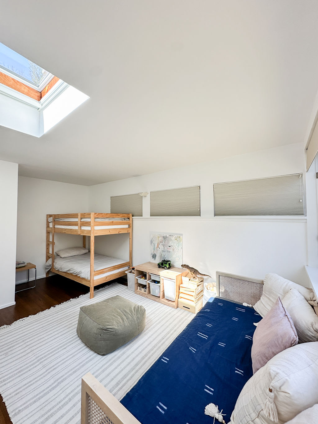 Bright kids' bedroom with a wooden bunk bed, a daybed, and a play area featuring a low wooden storage unit. A large skylight brightens the white-walled room, complemented by a striped area rug.