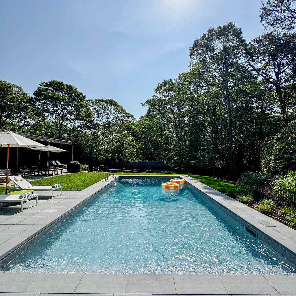 A long, rectangular swimming pool with clear blue water, bordered by grey paving stones and a manicured lawn, with lounge chairs and umbrellas on one side. Dense green trees line the perimeter under a bright blue sky.
