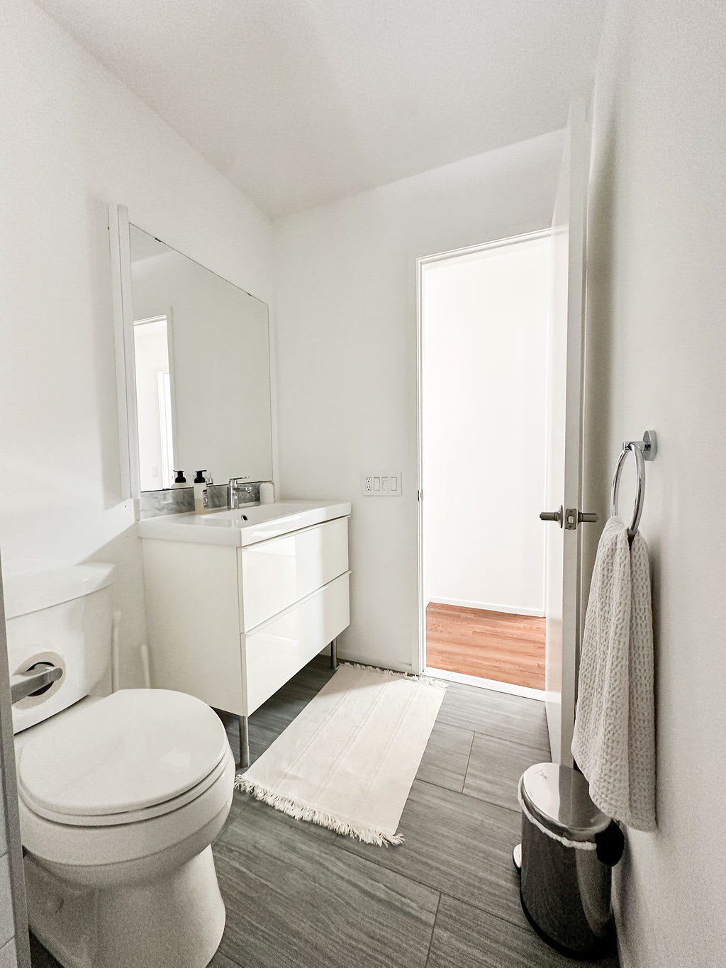 Modern white bathroom with a floating vanity, rectangular mirror, and a toilet. The floor is tiled in dark gray, and a light-colored rug adds warmth to the minimalist space.
