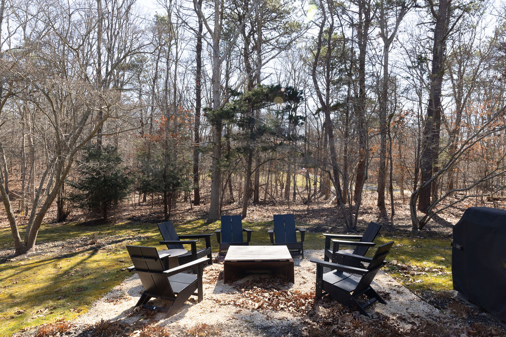 An outdoor gathering area with six black Adirondack chairs arranged around a low wooden coffee table, set on a gravel base. The area is surrounded by a dense, bare forest, suggesting a fire pit area.