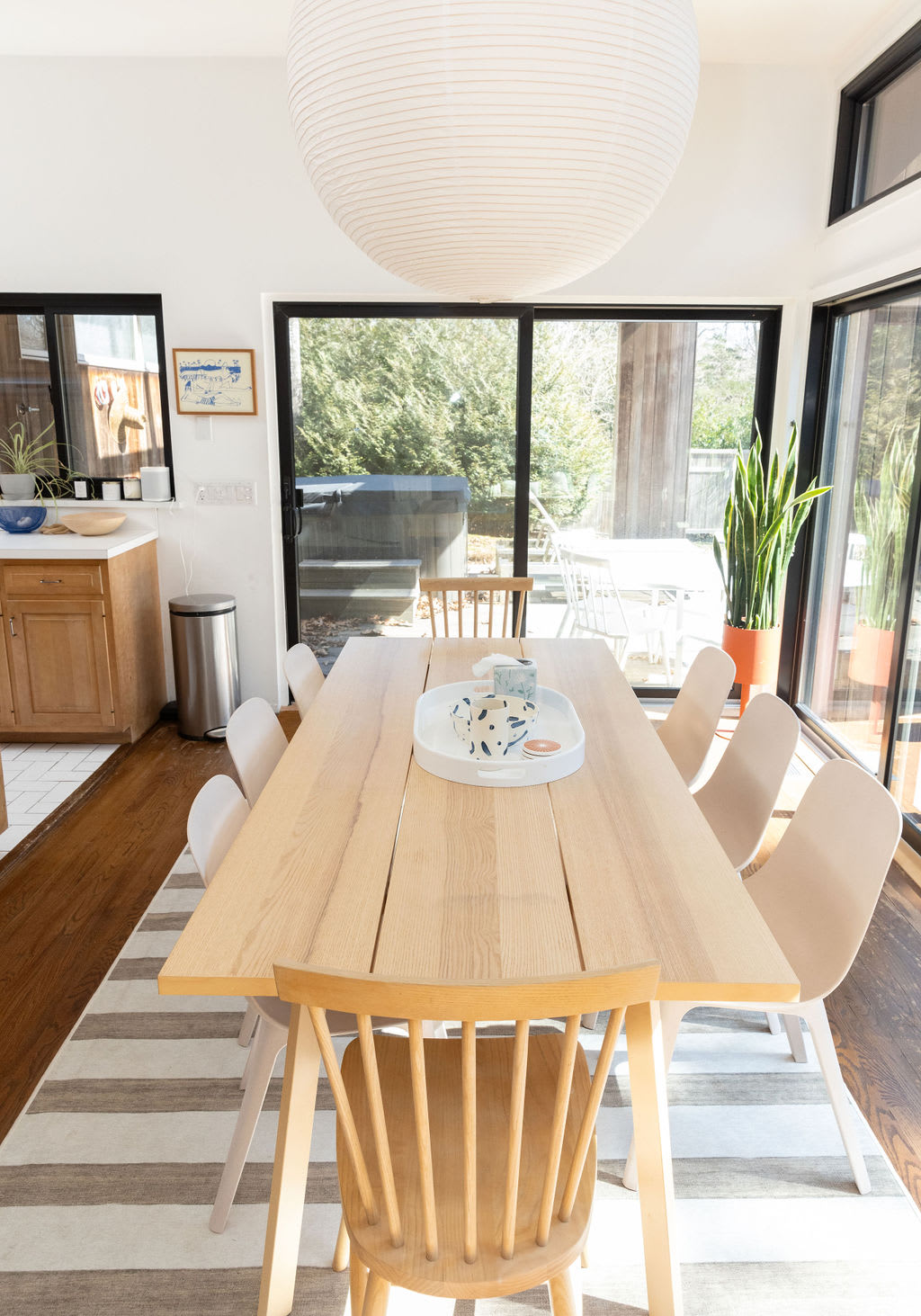 A close-up of the light wood dining table, elegantly set with a white tray and decorative items, surrounded by modern dining chairs, with a glimpse of the kitchen and outdoor patio through large windows.