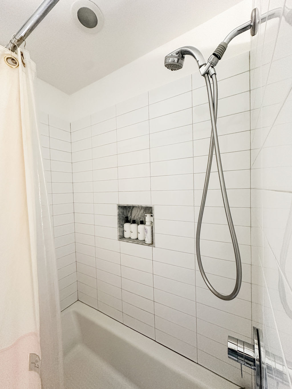 Close-up of a shower/tub combo with white subway tile walls and a built-in marble niche for toiletries. A flexible handheld showerhead is installed, alongside a simple shower curtain.