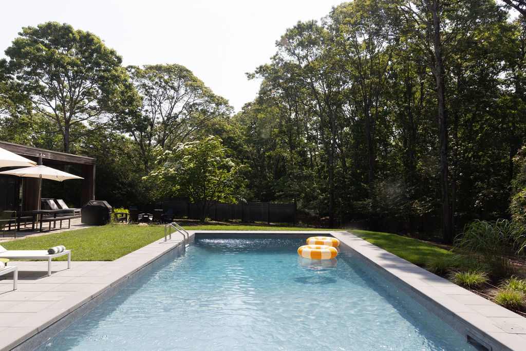 A clear blue rectangular swimming pool with grey paver coping, set in a grassy backyard surrounded by mature trees and a wooden fence. Two yellow and white inflatable rings float in the water.