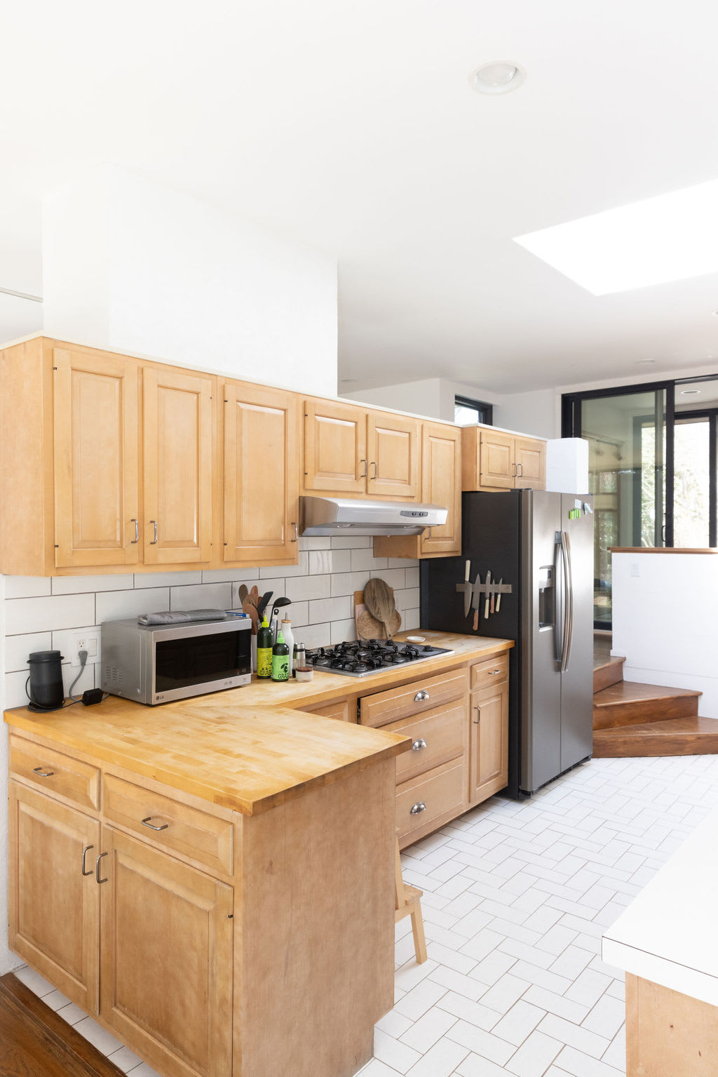 Wide shot of the kitchen showcasing light wood cabinets, butcher block countertops, and a white subway tile backsplash. It features a gas range, stainless steel refrigerator, and a small wooden step stool next to the island.