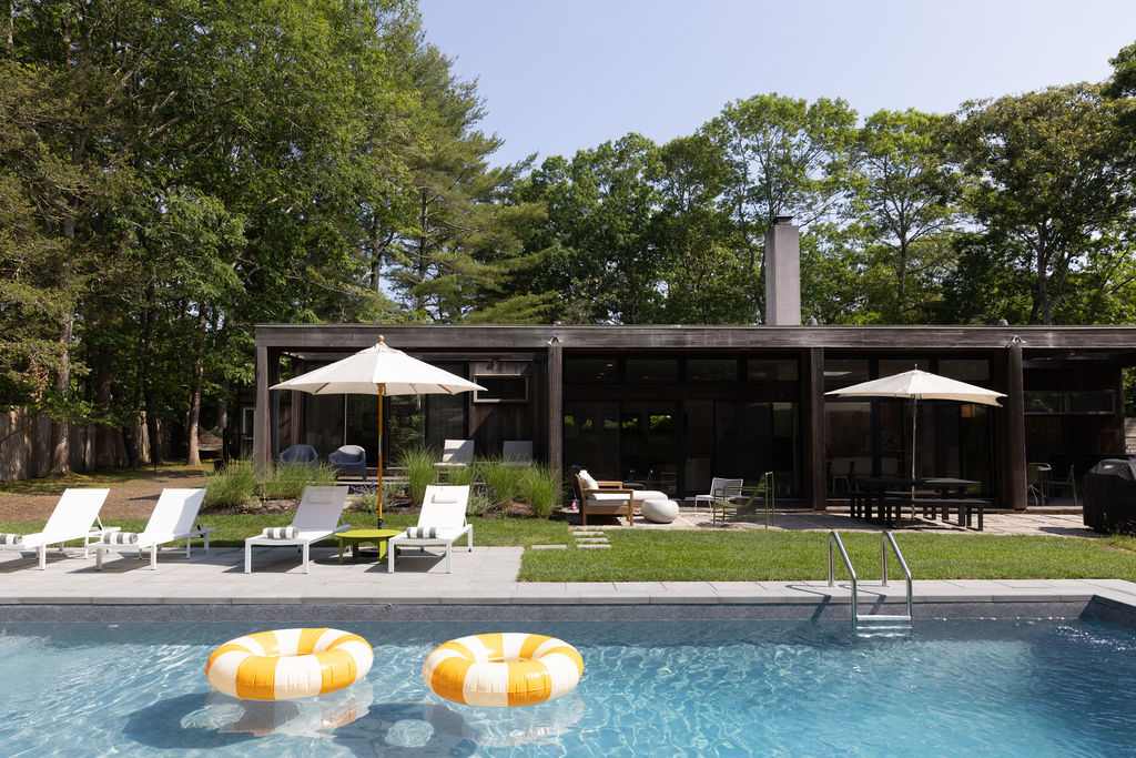 A modern house exterior with a rectangular swimming pool in the foreground, surrounded by a light-colored paved deck with white lounge chairs and umbrellas, set against a backdrop of lush green trees.