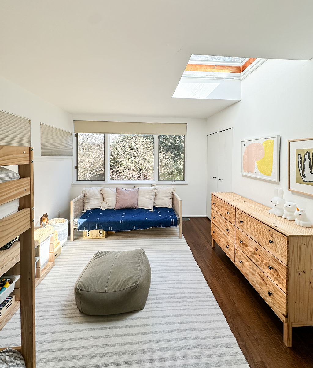 Spacious kids' bedroom featuring a wooden bunk bed, a daybed by a large window, and a long wooden dresser. A skylight and multiple windows provide ample natural light to the white-walled space.