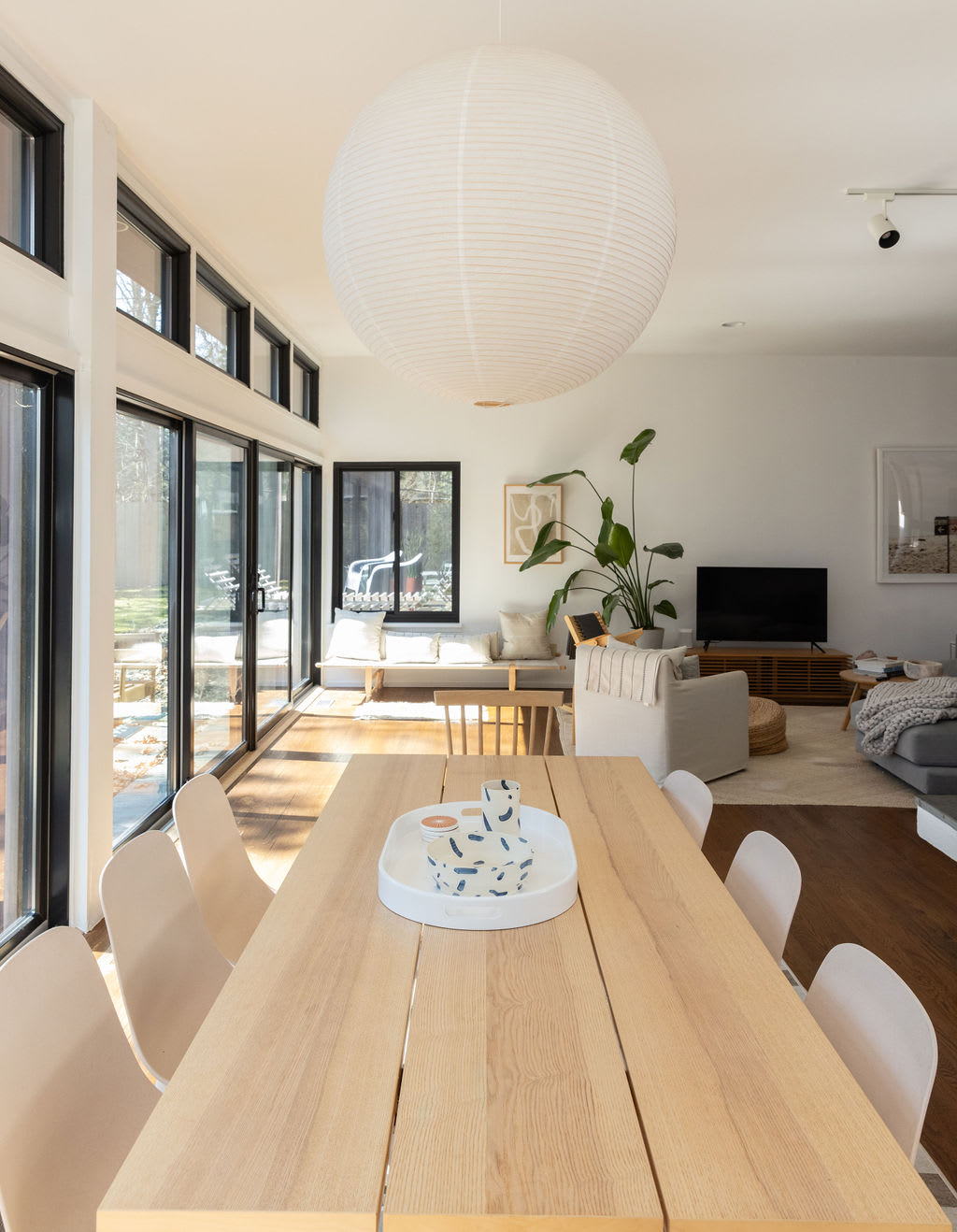 A spacious dining area with a large light wood table and chairs, illuminated by a prominent white paper lantern pendant light, offering a clear view into the adjacent living room and outdoor scenery through extensive windows.
