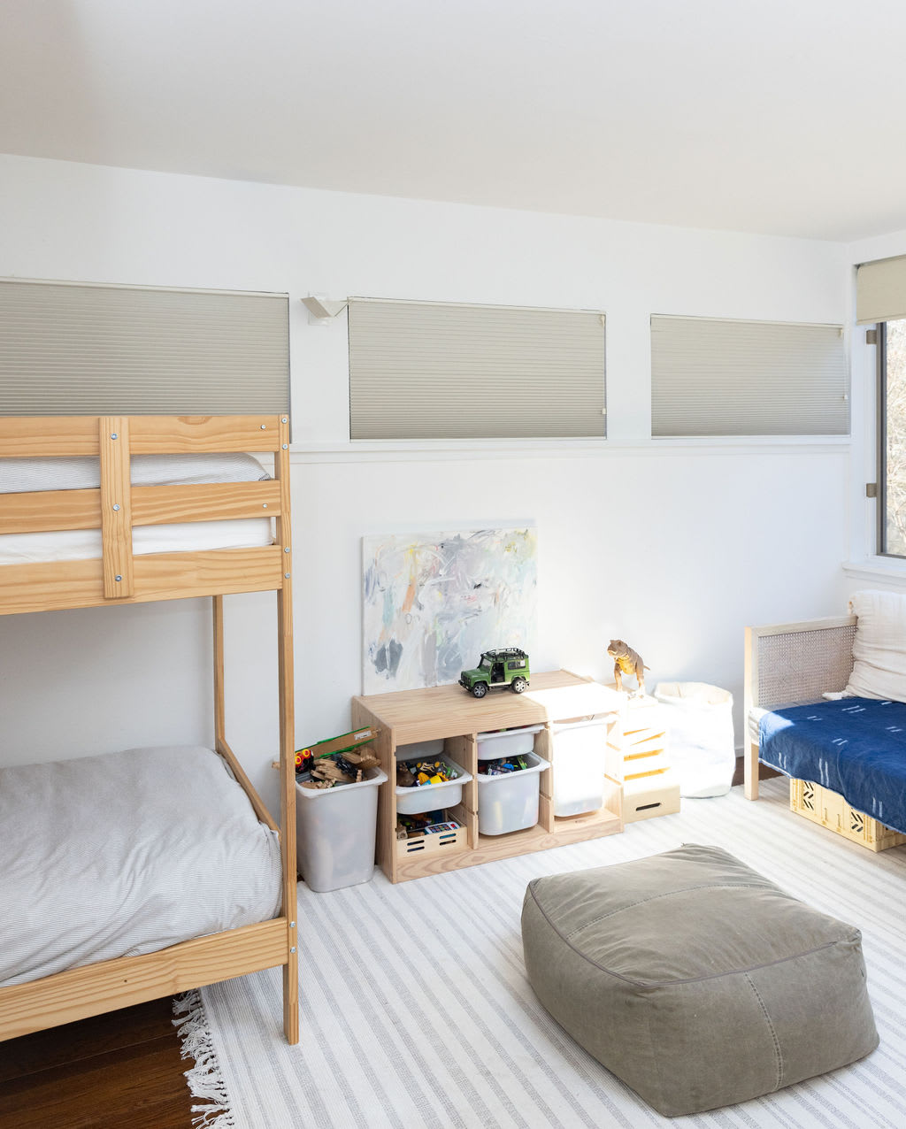 Kids' bedroom with a wooden bunk bed, a low wooden storage unit filled with toys, and a daybed. The room has white walls, light-colored blinds, and a striped area rug over dark wood floors.