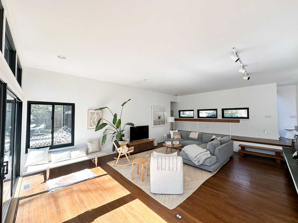 Spacious living area with hardwood floors, a large grey sectional, and a window bench with cushions. The room is brightly lit by numerous windows, including a row of transom windows, offering views of the lush outdoor surroundings.