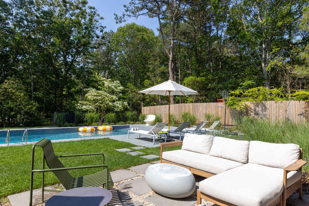 Inviting outdoor patio area with light-colored outdoor sofas and a green metal chair, set on square paver stones interspersed with gravel. The area overlooks the swimming pool and is surrounded by lush green grass and trees.