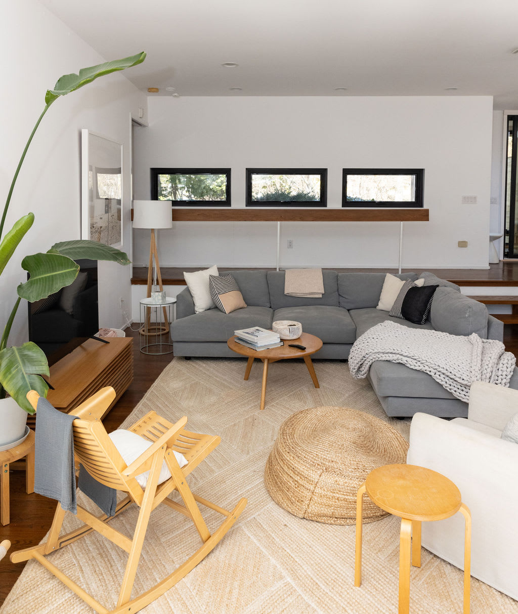 A spacious living room with a large grey sectional sofa adorned with throw pillows and blankets, centered around a round wooden coffee table on a light patterned rug, with a tall green plant adding a touch of nature.