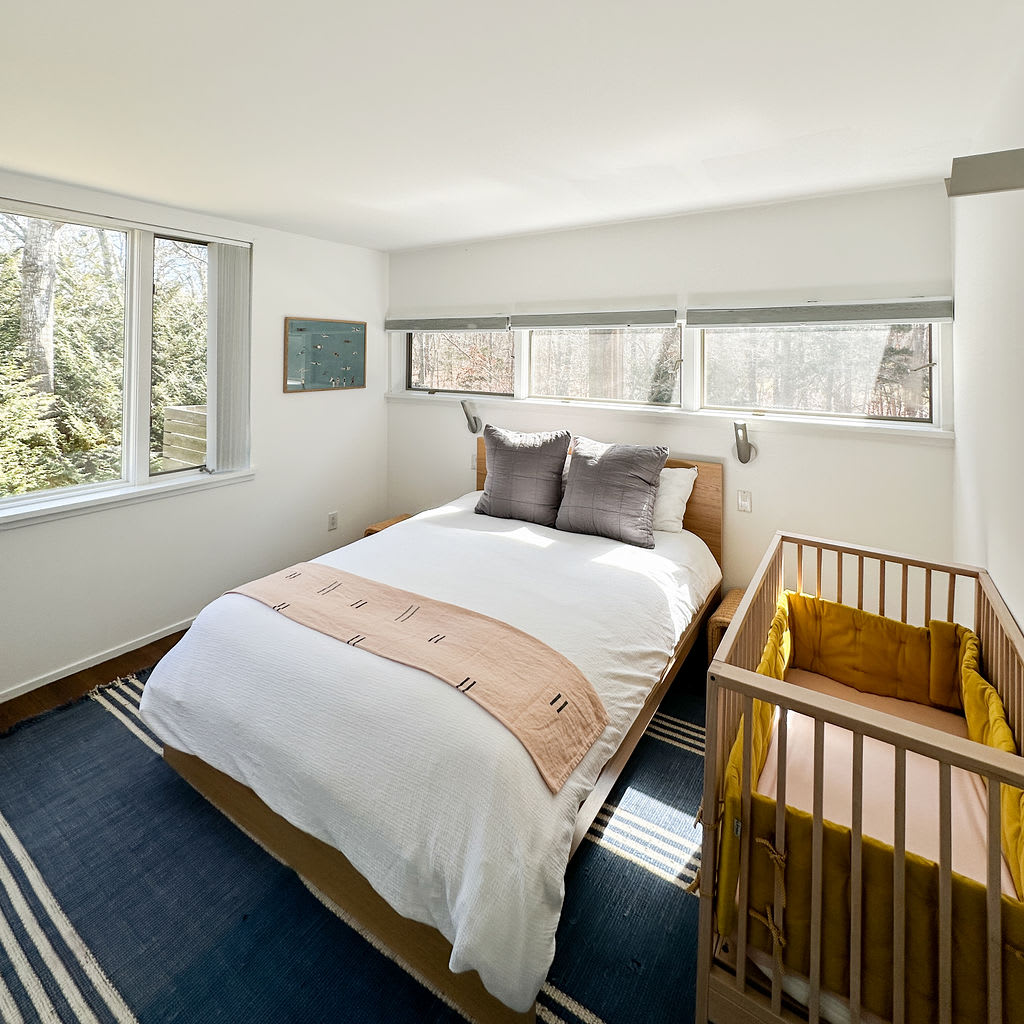 Angled view of the bedroom showing a light wood bed with white bedding, a blue striped rug, and a wooden crib. Large windows on the left and high horizontal windows above the bed provide ample natural light.