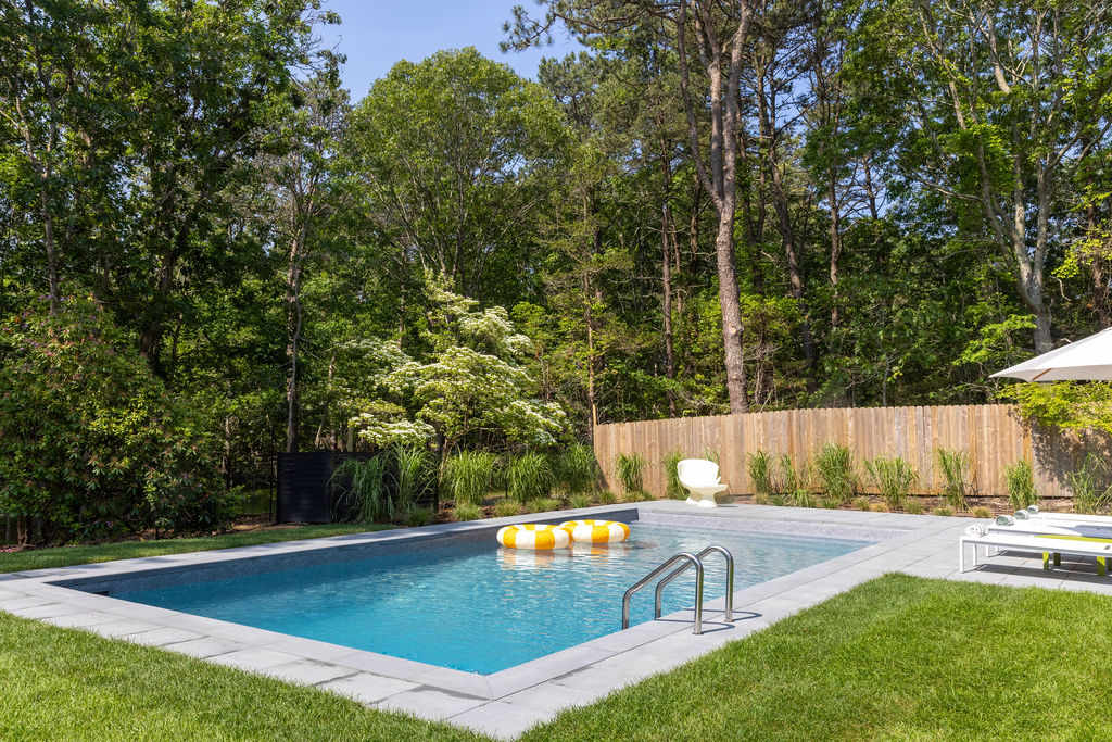 A rectangular swimming pool with light blue water, surrounded by grey paving stones and green grass, set against a backdrop of dense green trees. Two yellow and white striped inflatable rings float in the pool.