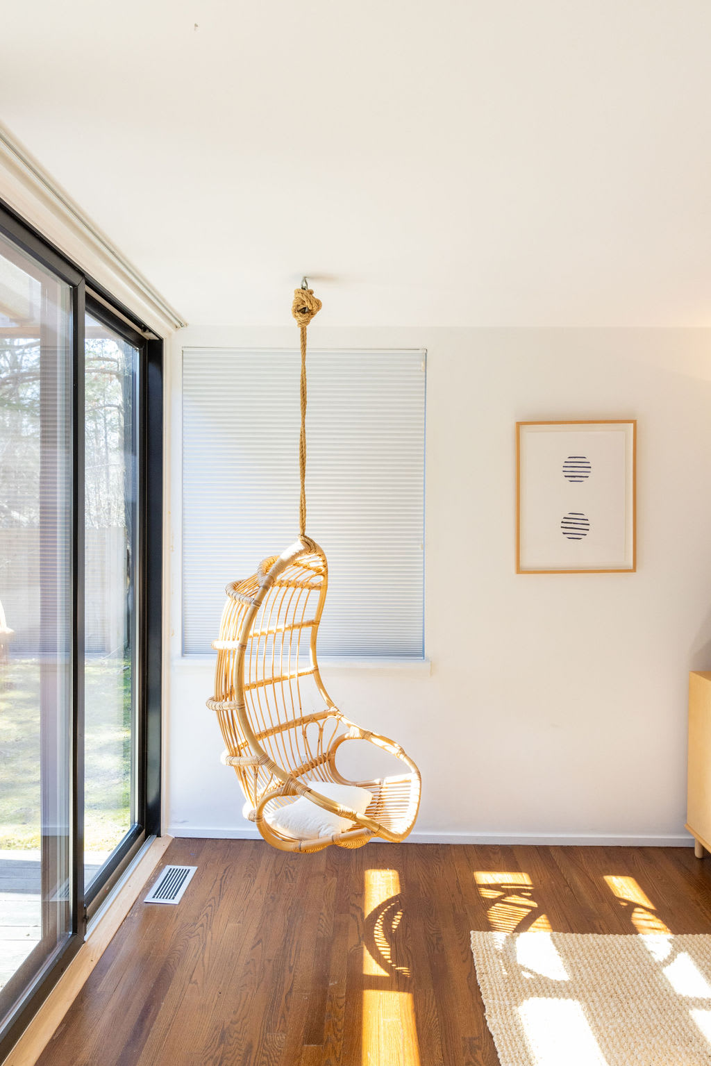 Bright room with dark wood floors and large sliding glass doors, featuring a hanging rattan egg chair with a white cushion. A framed abstract art piece adorns the white wall.