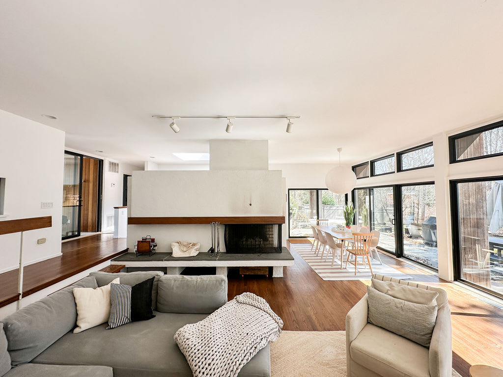 An open-concept living and dining area, showcasing a grey sectional sofa in the foreground, a central white fireplace, and a dining table with chairs visible through large windows in the background.