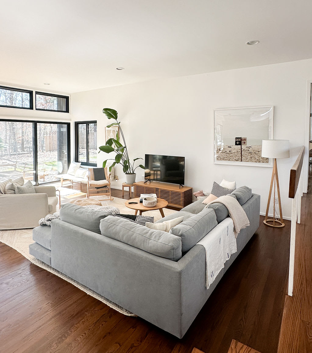 An expansive living room featuring dark hardwood floors, a large grey sectional sofa, a flat-screen TV on a wooden console, and floor-to-ceiling windows offering views of the surrounding outdoor greenery.