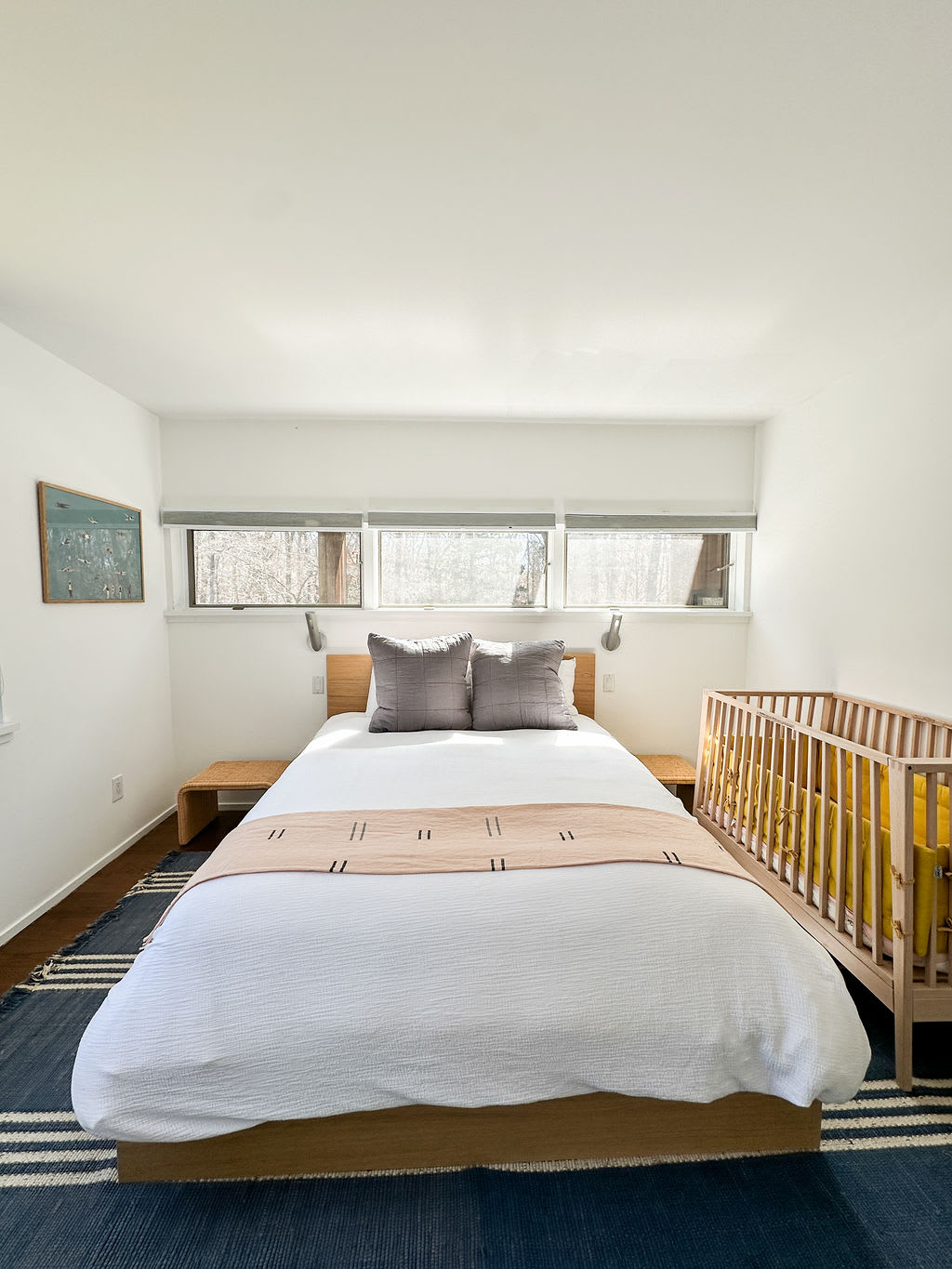 Wider shot of the bedroom with a light wood bed, white bedding, and a blue striped area rug. It features a wooden crib with yellow bedding next to the bed and high horizontal windows.