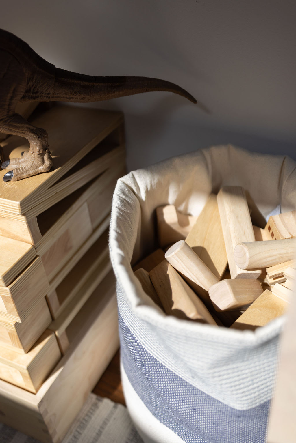 A close-up of a canvas bin filled with various wooden building blocks, next to a stack of wooden pallets and the tail of a dinosaur toy. The scene is dimly lit, suggesting an indoor play or storage area.